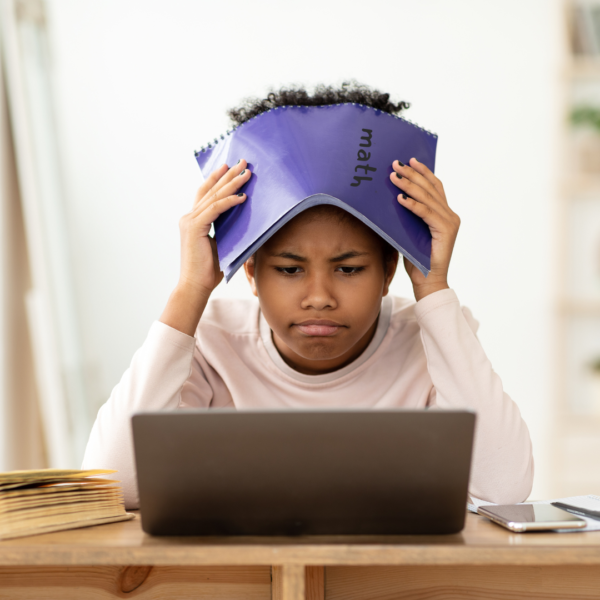 A child is holding a purple math notebook on his head. The student is looking at a school laptop and is frustrated with fractions. 